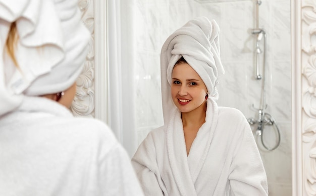 Mujer joven sonriente feliz en túnica blanca y con toalla en el baño en el espejo mirando a la cámara