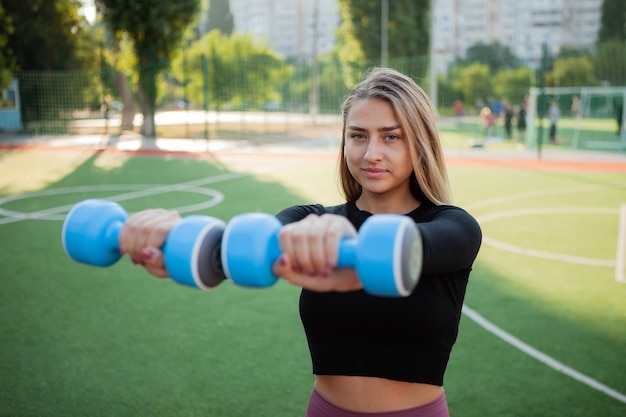 Mujer joven sonriente en forma practicando rizos con mancuernas delante de sí misma al aire libre