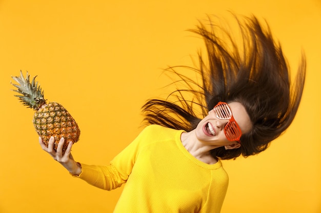 Mujer joven sonriente con gafas divertidas con el pelo volador sosteniendo fruta fresca de piña madura aislada en el fondo de la pared naranja amarilla. Estilo de vida vivo de la gente, concepto de vacaciones relajantes. Simulacros de espacio de copia