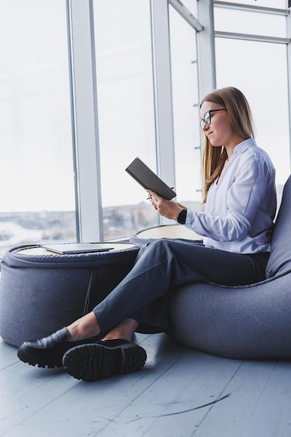 Mujer joven sonriente con gafas relajándose en una silla de bolsa de frijoles sentada en la mesa y relajándose usando el bloc de notas mujer feliz recostada disfrutando del trabajo sintiéndose satisfecha