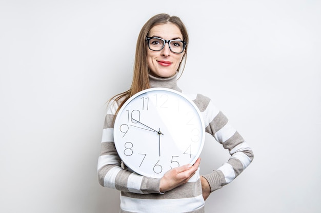 Mujer joven sonriente con gafas sosteniendo un reloj de pared blanco sobre un fondo claro