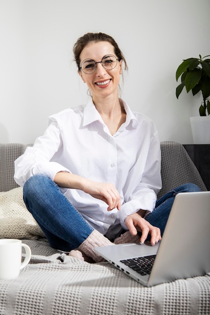 Mujer joven sonriente con gafas usando una laptop con café mientras se sienta en el sofá