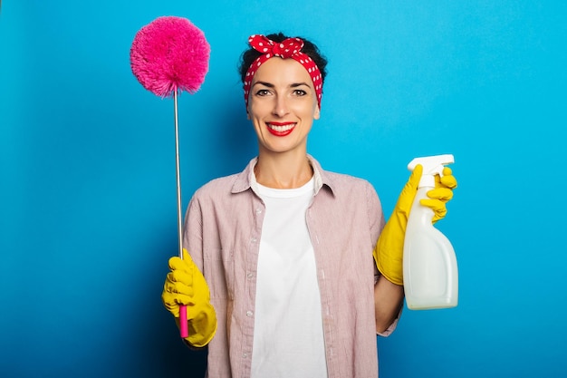 Mujer joven sonriente en guantes con spray y cepillo para limpiar el polvo sobre fondo azul.