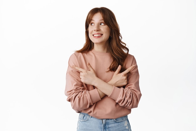 Mujer joven sonriente hace una elección, apuntando hacia los lados a la izquierda y a la derecha, comprando en la tienda, mirando los productos en la tienda, decidiendo, de pie en blanco