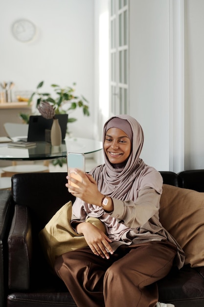 Mujer joven sonriente con hijab mirando la pantalla de su teléfono inteligente durante la comunicación en línea
