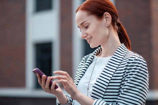 Mujer joven. Sonriente joven caucásica moderna en una chaqueta a rayas con su teléfono inteligente mientras está de pie al aire libre