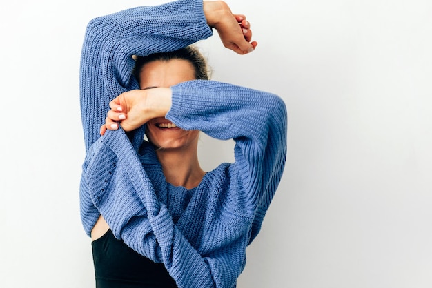 Mujer joven sonriente juguetona tirando de su suéter azul de moda sobre la cabeza divirtiéndose