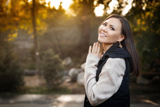 Mujer joven sonriente en el lado derecho de la foto se encuentra al aire libre en otoño con un suéter cálido y una chaqueta sin mangas