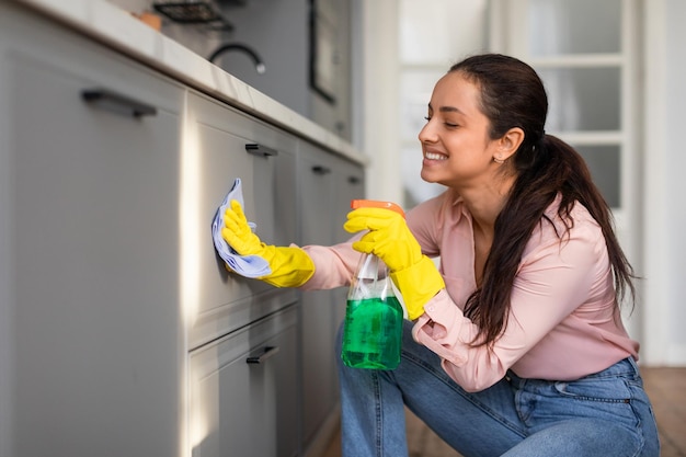 Mujer joven sonriente limpiando el gabinete de la cocina con spray y tela