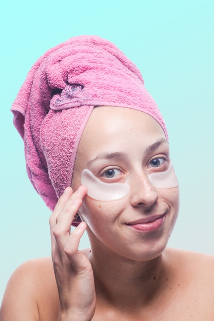 Mujer joven sonriente con las manchas blancas debajo de los ojos y una toalla rosada en su cabeza aislada en azul. Closeup retrato Protección de la piel