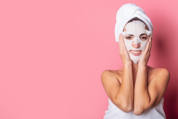 Mujer joven sonriente con una máscara cosmética en el rostro sobre un fondo rosa, tratamientos de spa