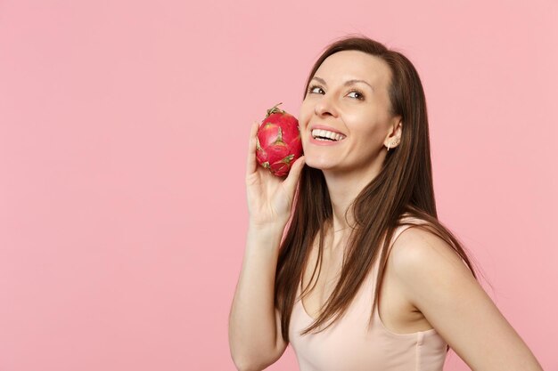 Mujer joven sonriente mirando hacia arriba sosteniendo en la mano pitahaya madura fresca, fruta de dragón aislada en el fondo de la pared rosa pastel en el estudio. El estilo de vida vívido de la gente relaja el concepto de vacaciones. Simulacros de espacio de copia.