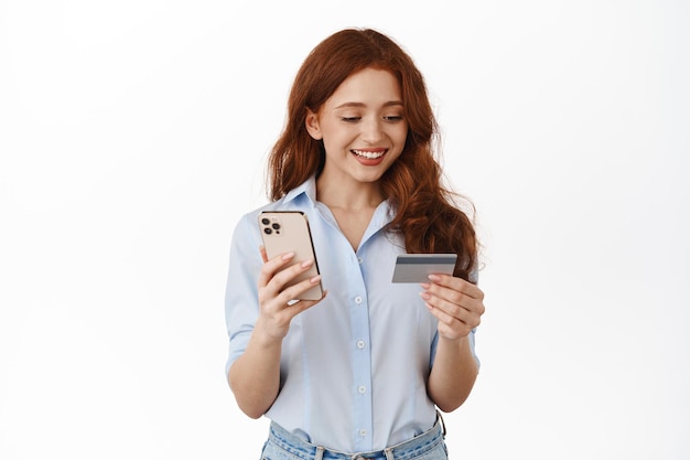 Mujer joven sonriente mirando la tarjeta de crédito, pagando con el teléfono móvil, ordenando en el sitio en Internet, comprando en línea, de pie contra el fondo blanco
