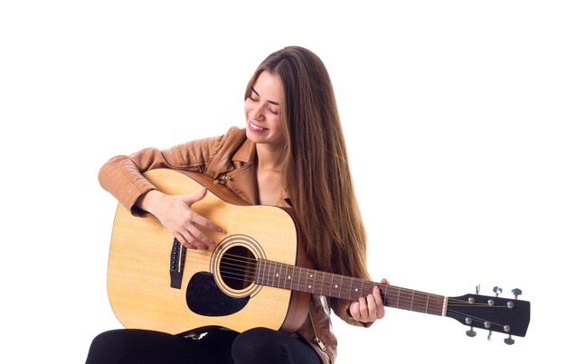 Mujer joven sonriente con el pelo largo en chaqueta marrón tocando la guitarra sobre fondo blanco en el estudio