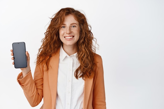 Mujer joven sonriente con el pelo rojo rizado que muestra la pantalla del teléfono móvil y se ve feliz demostrando la interfaz de la aplicación de pie contra el fondo blanco