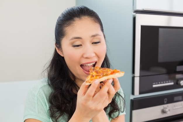 Mujer joven sonriente que come una rebanada de pizza en cocina