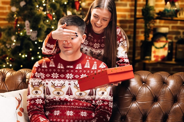 Mujer joven sonriente que cubre los ojos del marido curioso sonriente dando caja envuelta con regalo de Navidad