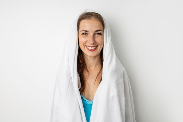 Mujer joven sonriente que cubre su cabeza con una sábana sobre un fondo blanco.