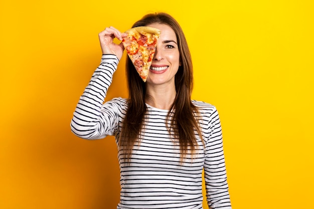 Mujer joven sonriente que cubre sus ojos con una rebanada de pizza fresca sobre un fondo amarillo