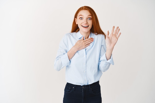 Foto mujer joven sonriente que mira con entusiasmo mientras hace una promesa, levanta una mano y hace un juramento, de pie sobre una pared blanca