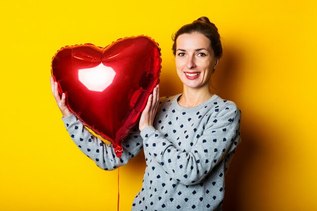 Mujer joven sonriente que sostiene el globo de aire en forma de corazón rojo sobre fondo amarillo.