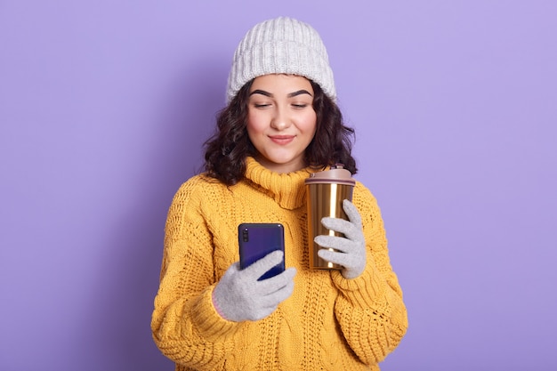 Mujer joven sonriente que sostiene el teléfono móvil y que bebe el café o el té caliente de la taza termo elegante