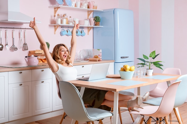 Mujer joven sonriente que usa la computadora portátil en la cocina en casa. Mujer rubia trabaja en computadora, freelance o blogger trabajando en casa