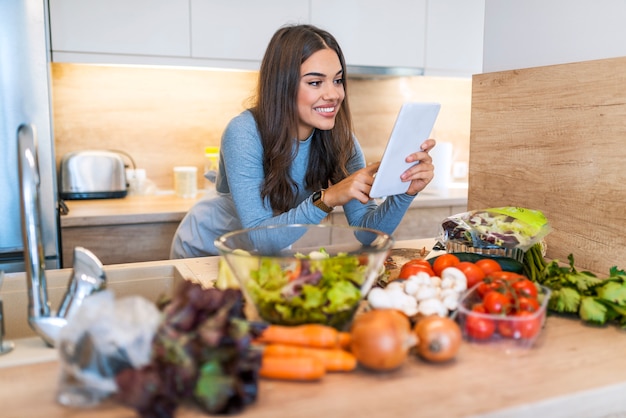 Foto mujer joven sonriente que usa la tableta de digitaces con la verdura en encimera en cocina