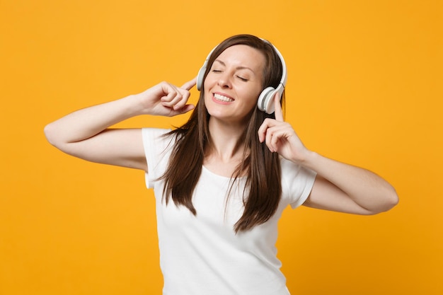 Mujer joven sonriente y relajada con ropa informal blanca que mantiene los ojos cerrados, escucha música con auriculares aislados en el fondo de la pared naranja amarilla en el estudio. Concepto de estilo de vida de las personas. Simulacros de espacio de copia.