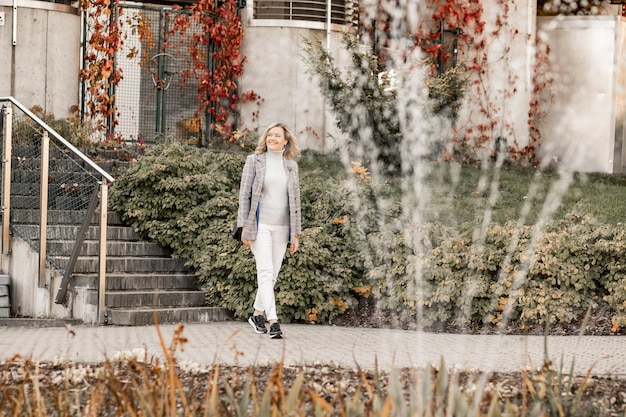 Mujer joven sonriente y relajada con suéter ligero, jeans blancos y chaqueta gris caminando a distancia al aire libre posiblemente en la ciudad o pueblo a principios de otoño con su hierba amarilla, hojas verdes y rojizas