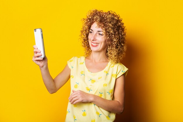 Mujer joven sonriente rizada hablando por un chat de video en el teléfono sobre un fondo amarillo.