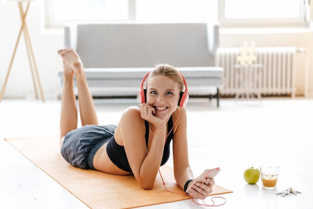 Mujer joven sonriente en ropa deportiva acostada en la alfombra del gimnasio escuchando música