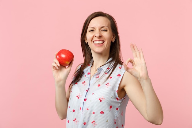 Mujer joven sonriente con ropa de verano que muestra un gesto OK sosteniendo fruta fresca de manzana roja madura aislada en un fondo de pared rosa pastel. Estilo de vida vivo de la gente, concepto de vacaciones relajantes. Simulacros de espacio de copia.