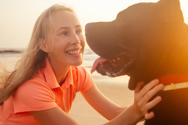 Mujer joven sonriente rubia abrazando a su perro Cane Corso en la playa