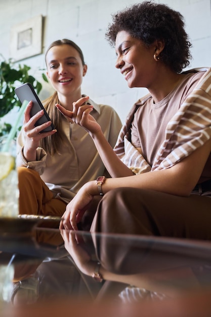 Mujer joven sonriente señalando la pantalla del teléfono inteligente sostenido por su amiga