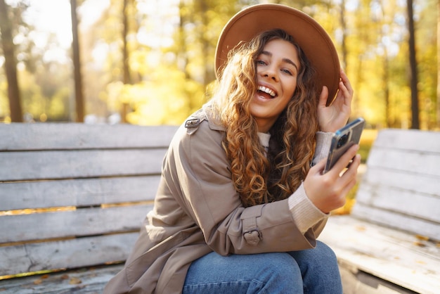 Mujer joven sonriente sentada en un banco en el parque de otoño usando el teléfono con ropa elegante de estado de ánimo feliz