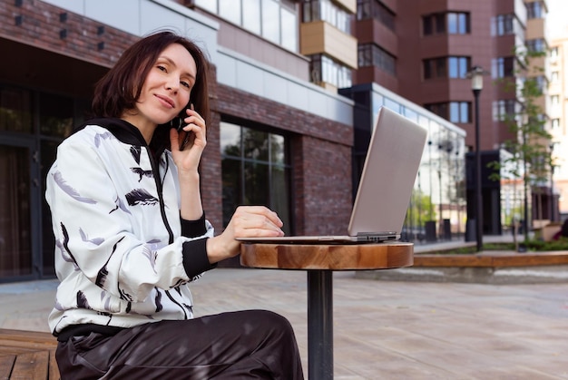 Mujer joven sonriente sentada en un café de la calle usando una computadora portátil y un teléfono Trabajando negocios en línea en línea