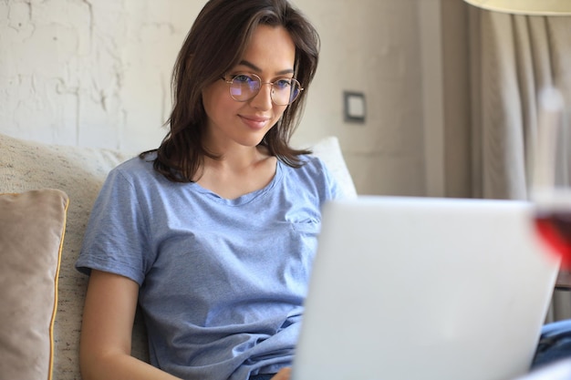 Mujer joven sonriente sentada en un sofá con computadora portátil y charlando con amigos