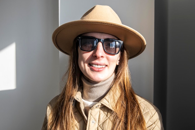 Mujer joven sonriente en un sombrero con gafas de sol contra una pared blanca