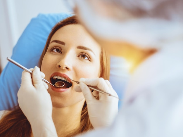 Mujer joven sonriente con soportes de ortodoncia examinados por un dentista en una clínica dental soleada. Dientes sanos y concepto de medicina.