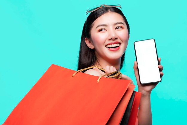 Mujer joven sonriente sosteniendo bolsas de compras y teléfono móvil en blanco contra un fondo azul