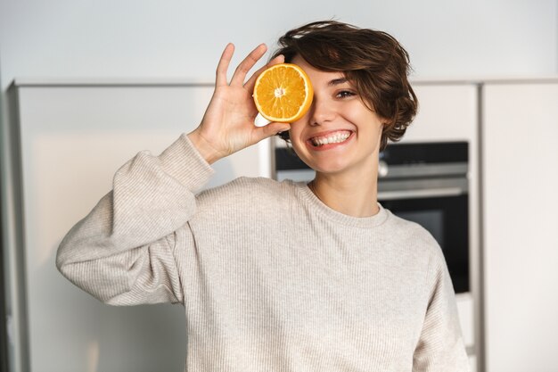 Mujer joven sonriente sosteniendo jugo de naranja y fruta de naranja mientras está de pie en la cocina