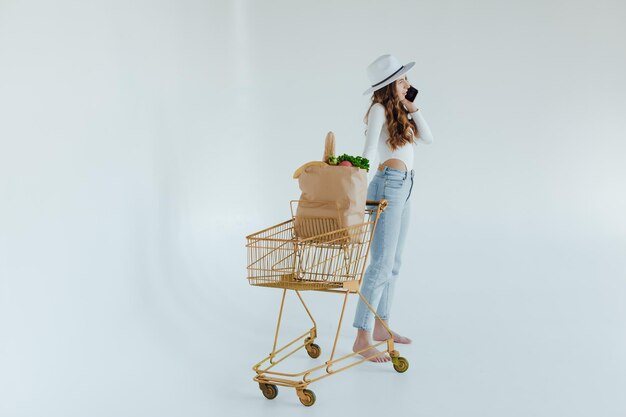 mujer joven sonriente sosteniendo manzana y mirando hacia otro lado mientras está de pie con carrito de compras