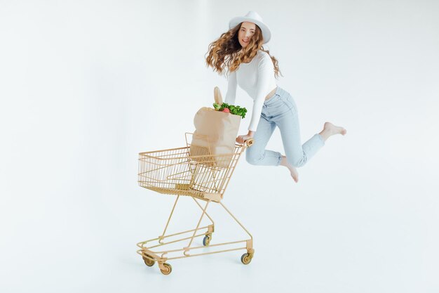 mujer joven sonriente sosteniendo manzana y mirando hacia otro lado mientras está de pie con carrito de compras