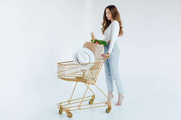 mujer joven sonriente sosteniendo manzana y mirando hacia otro lado mientras está de pie con carrito de compras