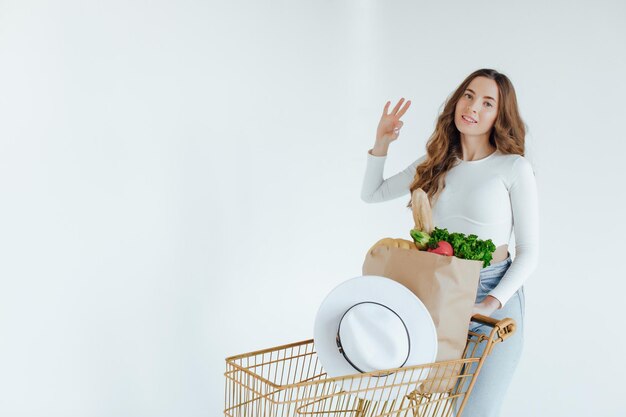 mujer joven sonriente sosteniendo manzana y mirando hacia otro lado mientras está de pie con carrito de compras