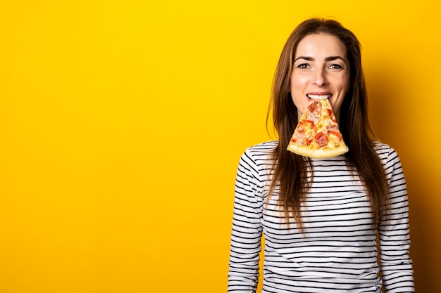 Mujer joven sonriente sosteniendo una rebanada de pizza fresca caliente en su boca en un amarillo.