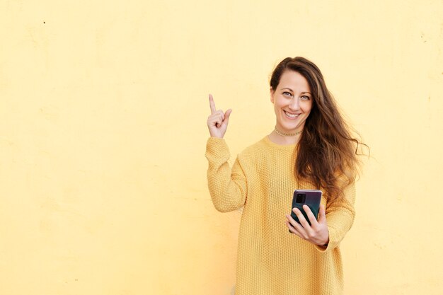 Mujer joven sonriente sostiene un teléfono celular con una pantalla vacía y señala su dedo índice hacia un lado