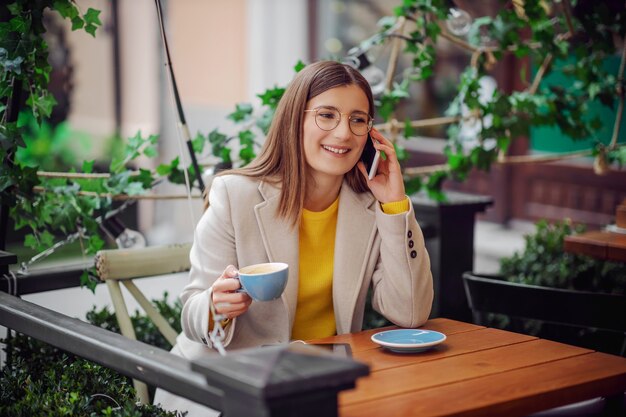 Mujer joven sonriente en suéter amarillo sentado en la terraza de la cafetería, sosteniendo una taza de café y hablando por un teléfono.