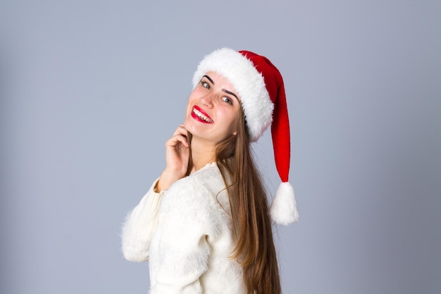 Mujer joven sonriente en suéter blanco con sombrero de navidad rojo sobre fondo gris en estudio